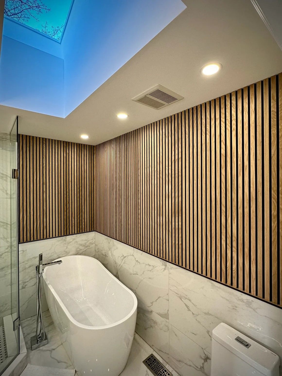 Bathroom with natural white oak slat panelling above marble tile, a freestanding tub and a skylight overhead.