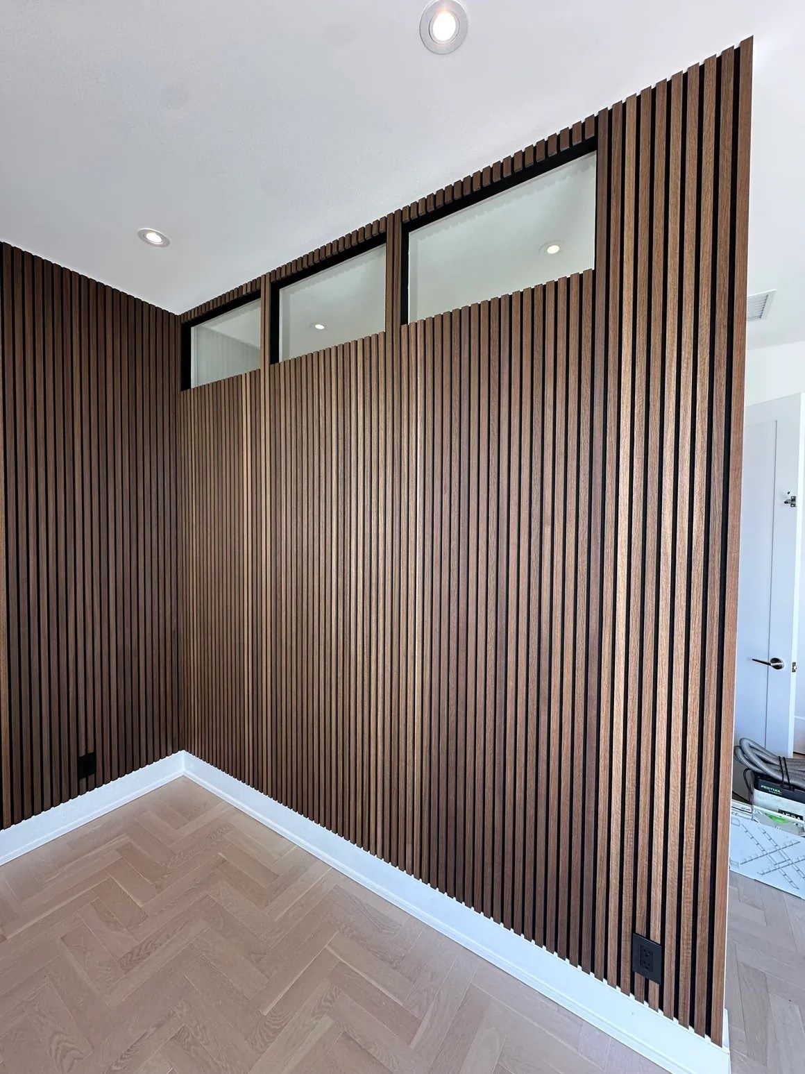 Oiled oak slats wrapping an interior corner and returning up to transom windows, above a herringbone floor.
