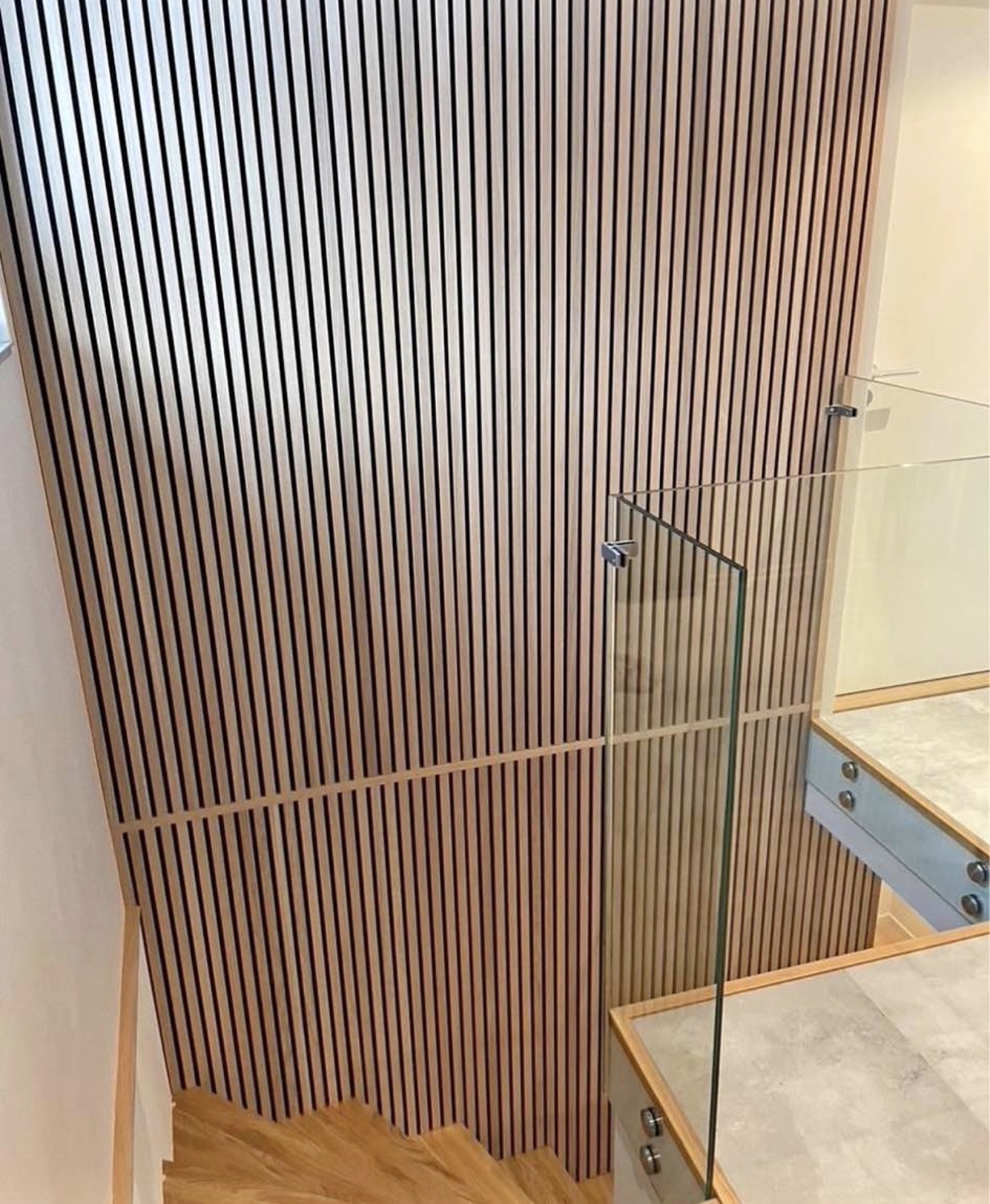 Stairwell landing with slats continuing from the wall up across the angled ceiling, beside a glass railing.